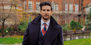 Man wearing a navy blue double breasted tweed long coat over a navy blue suit and a white shirt with a silk twill regimental tie with red and navy blue stripes and gold detailing