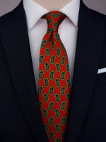Orange-red silk twill tie with a green and blue paisley pattern worn with a white shirt and navy blue suit