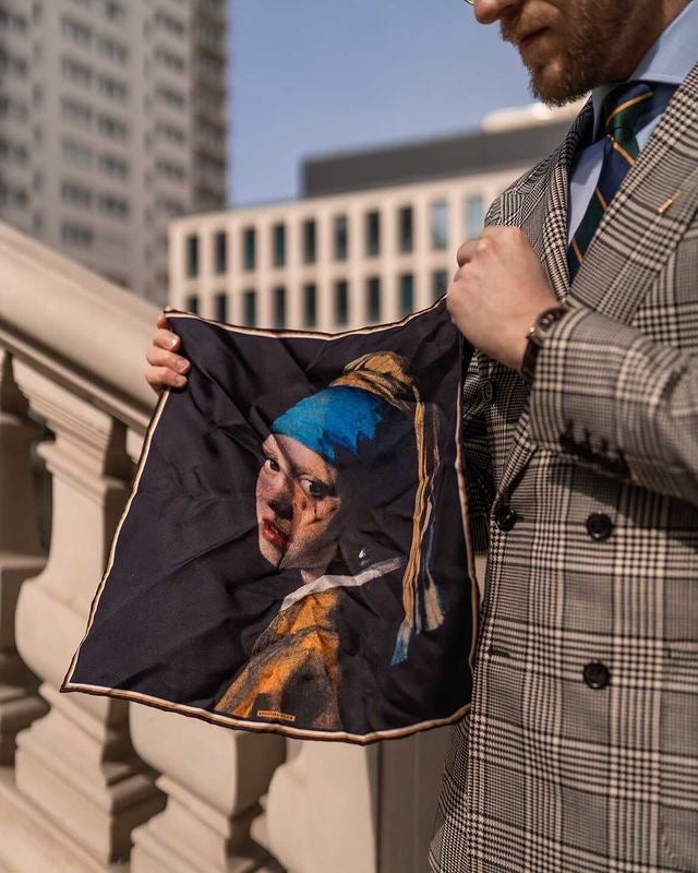 Man in a grey prince of Wales check suit wearing a green and navy blue striped regimental tie while holding a historical fine art silk pocket square of Vermeer's girl with a pearl earring. 