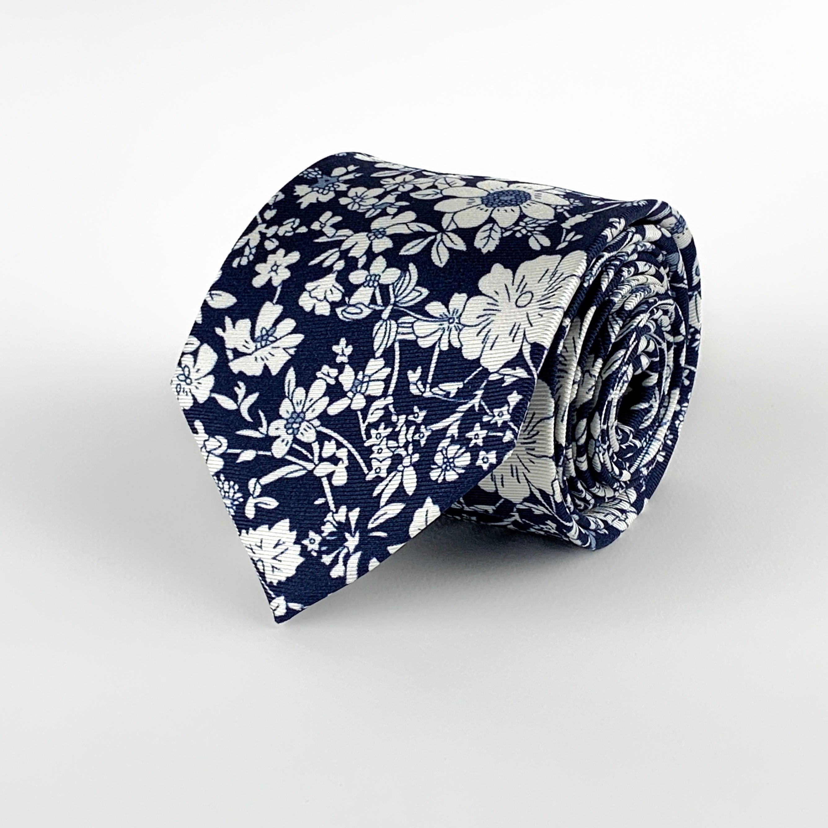 Navy blue mulberry silk twill tie with a white floral print rolled and placed on a white background