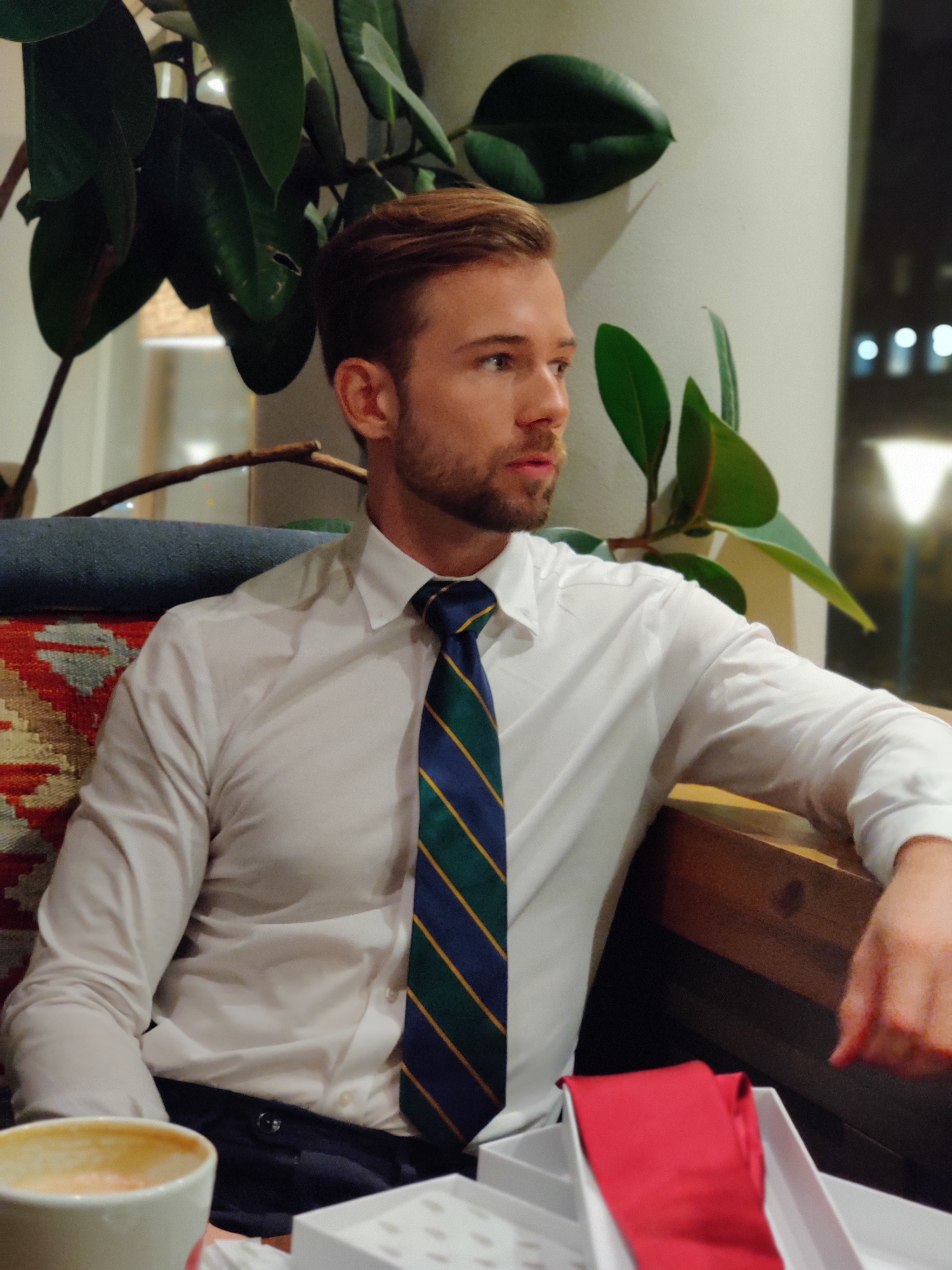 Man seated at a table at a cafe wearing a white shirt and a Mulberry silk twill tie with navy blue and dark emerald green regimental stripes and gold woven detailing