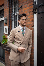 man leaning on a brick townhouse wall while wearing a beige double breasted suit with a white shirt, a Mulberry silk twill tie with a multi-colored rainforest floral print on a black base and glasses. 