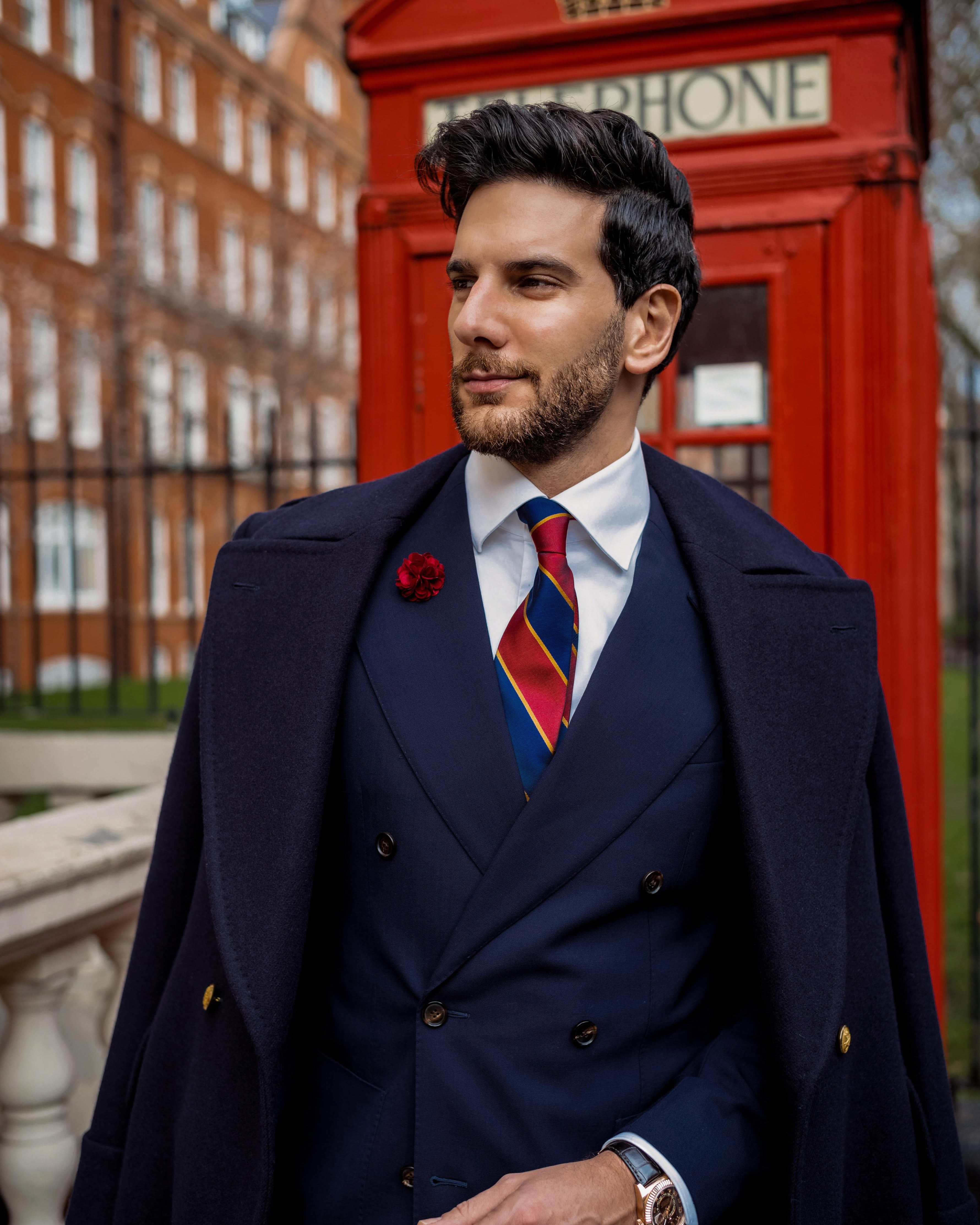 man standing in front of a red telephone booth in London wearing a navy blue long coat and a navy blue double breasted suit with a white shirt and a red and navy blue striped regimental mulberry silk twill tie with gold woven detailing