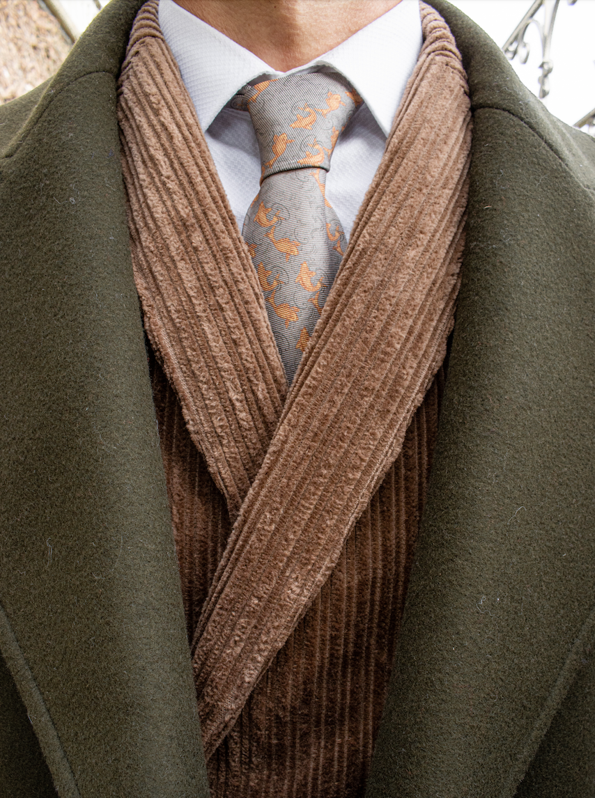 Man wearing a green tweed winter coat with a textured suede velvet double breasted jacket underneath paired with a white shirt and a Mulberry silk twill tie in a grey color with a printed pattern of orange fish placed in diagonal stripes