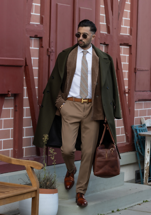 Man standing next to a wall  wearing a green tweed winter coat with a textured suede velvet double breasted jacket underneath paired with a white shirt and a Mulberry silk twill tie in a grey color with a printed pattern of orange fish placed in diagonal stripes. He is also wearing a brown formal pant with a Hermes belt and brown leather shoes while carrying a brown leather bag